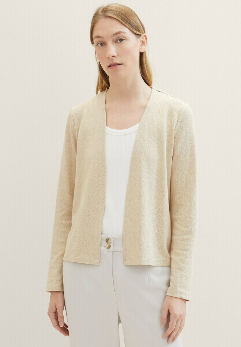 Beige cardigan med avslappnad passform, öppen front och långa ärmar. Tillverkad av ett texturerat stickat tyg. Matchad med en vit topp och ljusa byxor.