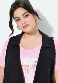 Schwarze Weste über einem rosa T-Shirt mit einem glänzenden, bunten "Happy"-Grafik, mit kurzen Ärmeln und einem dezenten V-Ausschnitt.