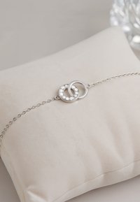 Silverarmband på en mjuk, blek kudde. Har två sammanlänkade cirkulära länkar, varav en är prydd med klara ädelstenar och har en slät yta.