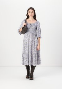 Rochie maxi cu imprimeu floral în nuanțe de violet și alb, având un corp smockat și fustă în straturi. Asortată cu o geantă neagră și cizme maro până la genunchi.