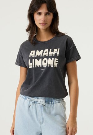 Schwarzes Baumwoll-T-Shirt mit umgeschlagenen Ärmeln, auf dem in weißer, fetter Schrift "AMALFI LIMONE" steht. Kombiniert mit hellblauen, mit Kordelzug versehenen Hosen.