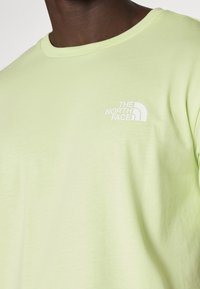 Limetkově zelené tričko vyrobené z měkké tkaniny, s kulatým výstřihem a malým bílým logem na levé části hrudi. Hladká textura a neformální design.