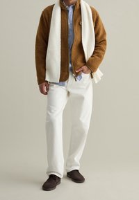 Bruine fleecetrui met een ritssluiting, witte geribbelde sjaal, gestreept blauw overhemd, witte broek en donkerbruine schoenen, staande op een neutrale achtergrond.