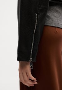 Casaco de couro preto com punhos com fecho de correr, combinado com uma blusa cinza e uma saia castanha. O casaco apresenta uma textura suave e um design elegante.