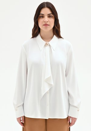 Femme aux longs cheveux foncés portant un chemisier blanc avec une broche décorative au col et un pantalon large marron, debout devant un fond blanc.