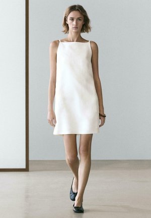 Ung kvinde, der går indendørs iført en ærmeløs hvid minikjole og sorte ballerinasko, mod en ensartet grå væg som baggrund.