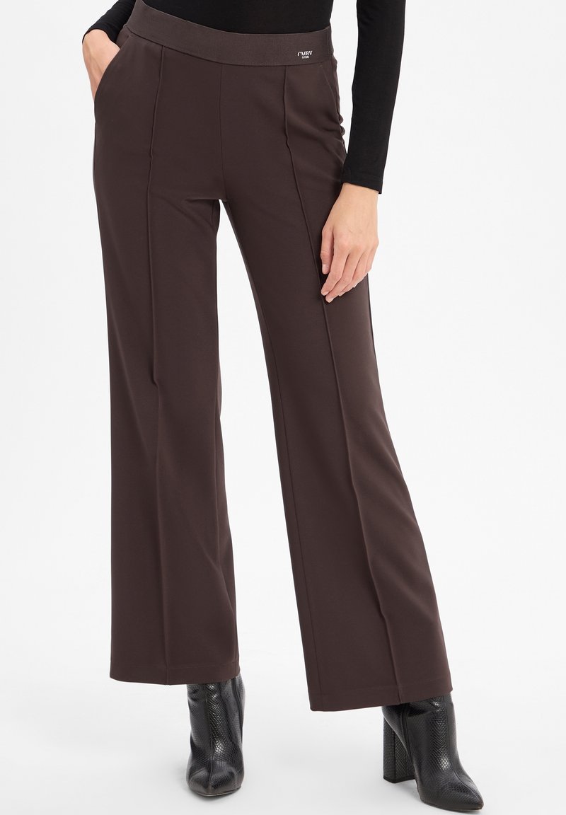 Femme portant un pantalon marron taille haute à jambes larges avec des coutures devant, associé à des bottines noires à talons et un top noir à manches longues.