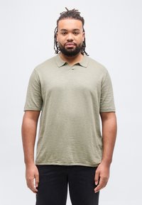 Νέος άντρας με κοντά dreadlocks, φοράει ανοιχτόχρωμο πουκάμισο πόλο σε ελιά πράσινο και μαύρο παντελόνι, στέκεται μπροστά σε λευκό φόντο.