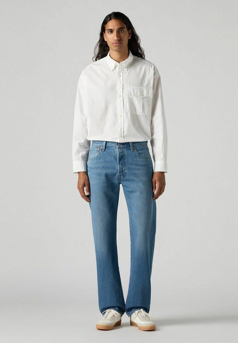 Chemise blanche à boutons avec une poche poitrine, associée à un jean en denim bleu clair et des baskets blanches avec une semelle en caoutchouc beige.