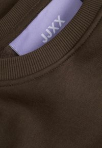 Bruine sweatshirt met een geribbelde halslijn en een lichtpaarse label met de tekst "JJXX." De stof lijkt zacht en heeft een textuur.