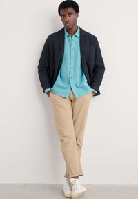 Blazer bleumarin, cămașă cu nasturi bleu deschis, chinos bej și teniși albi. Materialele par a fi fine și bine ajustate, cu mânecile suflecate.