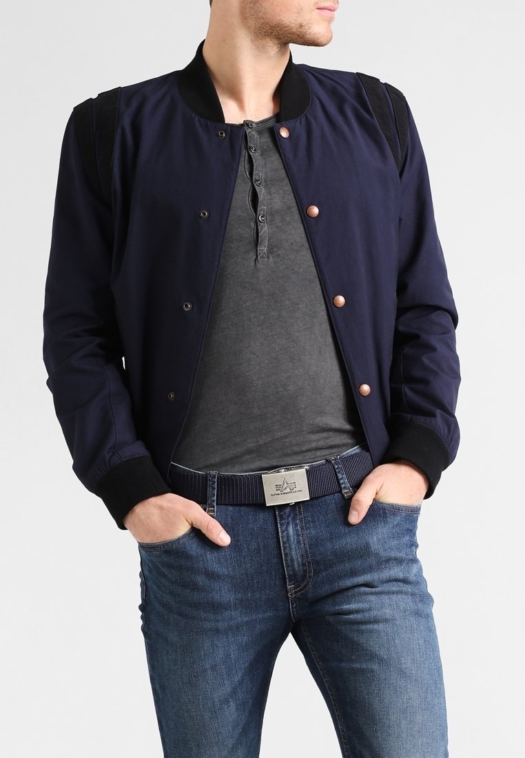 Homme portant une veste bomber bleu marine par-dessus un t-shirt henley gris foncé, jean bleu et ceinture bleue marine avec boucle argentée, mains dans les poches.