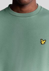 Uomo che indossa un maglione girocollo verde salvia con un piccolo logo di uccello giallo sul lato sinistro del petto.