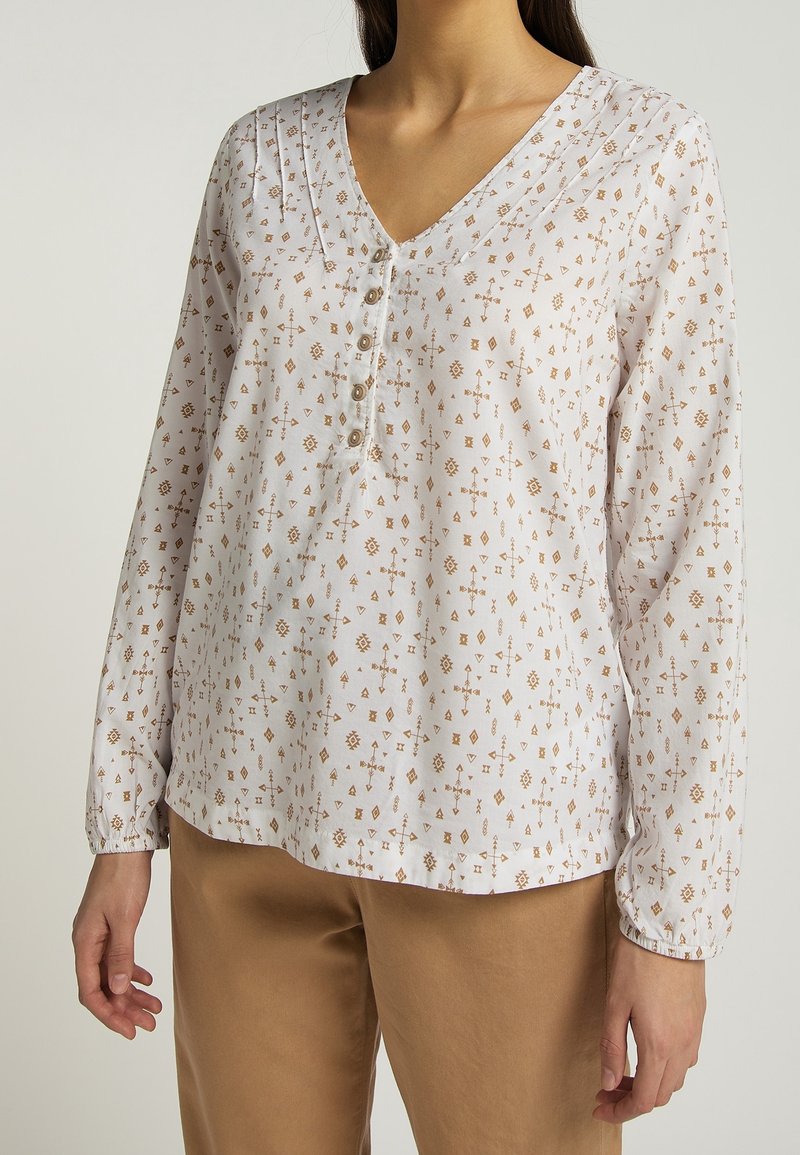 Femme portant un chemisier blanc à manches longues avec des imprimés géométriques bruns et des boutons, assorti à un pantalon beige, sur un fond uni.