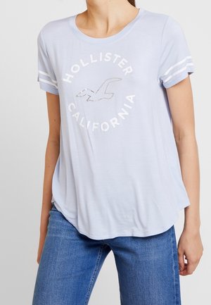 Frau trägt ein hellblaues Hollister California T-Shirt mit weißen Streifen an den Ärmeln und blaue Jeans, steht vor einfachem Hintergrund.
