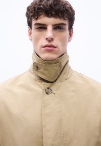 Νεαρός άνδρας με σγουρά μαλλιά που φοράει μπεζ παλτό με ψηλό γιακά κουμπωμένο στο λαιμό, κοιτάζοντας προς τα εμπρός με φόντο λευκό και απλό.