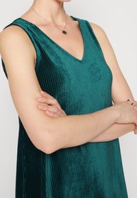 Teal ribbattu velvetipuku, jossa on V-muotoinen kaula-aukko ja hihaton muotoilu, näyttää pehmeältä ja väljältä. Näkyvissä on hopeaketju.