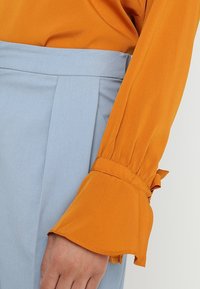 Oranssi pitkähihainen paita, jossa on rypytetyt hihansuut, yhdistettynä vaaleansinisiin korkeavyötäröisiin housuihin, joissa on sileä pinta ja taitteet.