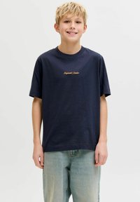 Χαμογελαστός ξανθός αγόρι που φοράει μπλε ναυτικό μπλουζάκι με πορτοκαλί κείμενο "Originals Studio" και ανοιχτό μπλε τζιν, στέκεται μπροστά σε απλό φωτεινό φόντο.