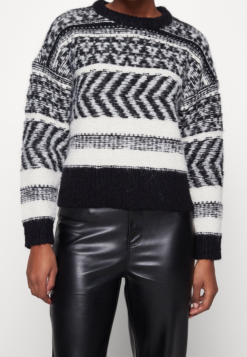 Pull en maille à motifs noir et blanc porté avec un pantalon en cuir noir, montrant le torse et les bras sur un fond neutre.