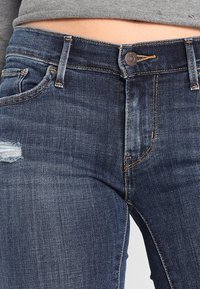 Person wearing dark blue denim jeans with a small distressed tear on the left thigh and a gray cropped top.