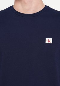 Tmavomodré bavlnené tričko s okrúhlym výstrihom a krátkymi rukávmi. Na tričku je malá obdĺžniková biela nášivka s červenými detailmi.