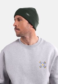 Bonnet côtelé vert foncé avec une finition texturée. Porté avec un sweat-shirt gris clair ornée d'une broderie florale en bleu, jaune et vert.