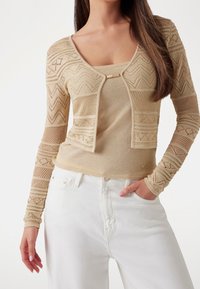 Beige neulottu neuletakki, jossa on kuvioitu design, avoin etuosa ja pitkät hihat, yhdistettynä vastaavaan toppiin ja valkoisiin farkkuihin.