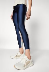Granatowe legginsy o błyszczącej powierzchni, z pastelowymi panelami bocznymi. Białe buty sportowe z teksturowanym materiałem i jasnymi akcentami.