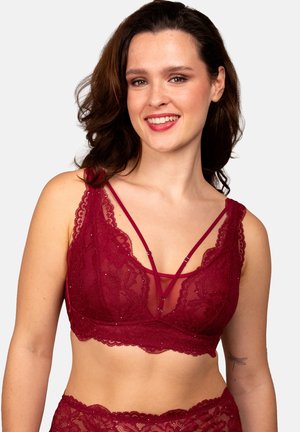 Lächelnde Frau mit welligem braunem Haar, die ein rotes Spitzen-Bralette mit dünnen Trägern und passende Spitzenunterwäsche vor einem schlichten Hintergrund trägt.