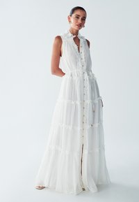 Abito maxi bianco con colletto arricciato, chiusura a bottoni e design a strati. Realizzato in tessuto testurizzato con motivi sottili e una vita annodata.