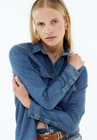 Denim shirt, light blue, long sleeves, buttoned cuffs, two chest pockets, soft texture, worn with a brown belt, tucked into high-waisted jeans.
