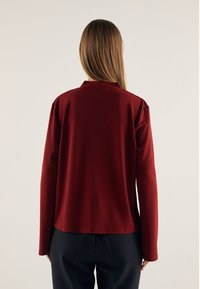 Bluză lungă cu mâneci lungi, de culoare burgundă, cu textură moale, având un guler discret și croială lejeră. Asortată cu pantaloni închiși la culoare.