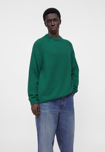 Homme portant un pull en maille vert ample et un jean bleu taille décontractée, debout devant un fond gris uni.
