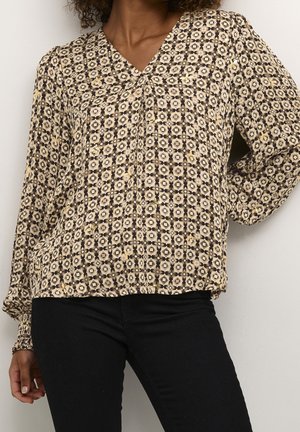 Mujer con blusa de manga larga de patrón geométrico en beige y negro con escote en V y pantalones negros ajustados, posando con una mano en la cadera.