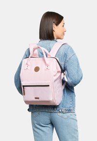 Roze rugzak met een rechthoekige vorm, stoffen materiaal, een voorste ritszak, twee zijvakjes en verstelbare schouderbanden. Bruine logodetail.