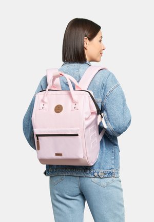 Pink backpack with a rectangular shape, fabric material, front zip pocket, two side pockets, and adjustable straps. Brown logo detail.