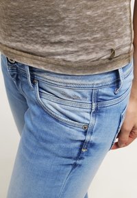 Světle modré džíny s úzkým střihem, s kontrastním prošitím, předními kapsami a malou značkovou etikettou blízko pasu.