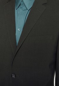 Schwarze Sakkajacke mit Reverskragen, Ein-Knopf-Verschluss und einer Brusttasche. Darunter ist ein hellblaues Hemd mit Knopfdetails sichtbar.