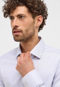 Camicia da uomo di colore viola chiaro con texture, colletto alla francese e polsi con bottone. Il modello sistema il colletto con una mano, mostrando uno stile informale.