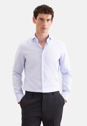 Homme aux cheveux bouclés portant une chemise habillée bleu clair boutonnée et un pantalon sombre, debout les mains dans les poches sur fond blanc.