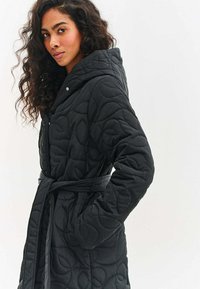 Žena s kučeravými vlasmi, oblečená v čiernom prešívanom kabáte so stuhou a kapucňou, stojaca na svetlom, jednoliatom pozadí, mierne otočená nabok.