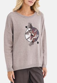Beżowy sweter z dzianiny z okrągłą grafiką przedstawiającą stylizowane dłonie i okrągłe akcenty. Posiada okrągły dekolt i ściągacze na rękawach.