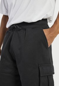 Personne portant un pantalon cargo noir avec une taille élastique et un t-shirt blanc, la main reposant dans la poche latérale.