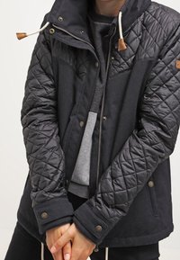 Veste noire à texture matelassée, dotée d'une capuche amovible, d'une fermeture éclair et d'accents en cuir marron sur les boutons-pression et le logo.