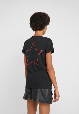 Persona con pelo corto y rizado, vistiendo una camiseta negra con el contorno de una estrella roja en la espalda y pantalones cortos de cuero negro, de espaldas.
