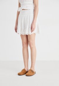 Jupe skater blanche brodée avec un ourlet festonné et une taille élastique, associée à des sabots en suede beige avec un détail de boucle.