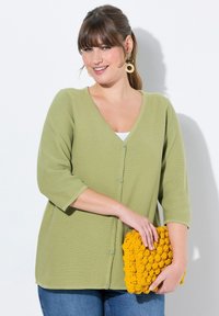 Olivgrön kofta med V-ringning och trekvartsärmar, med texturerat tyg och knappar, i kombination med en gul virkad clutch.