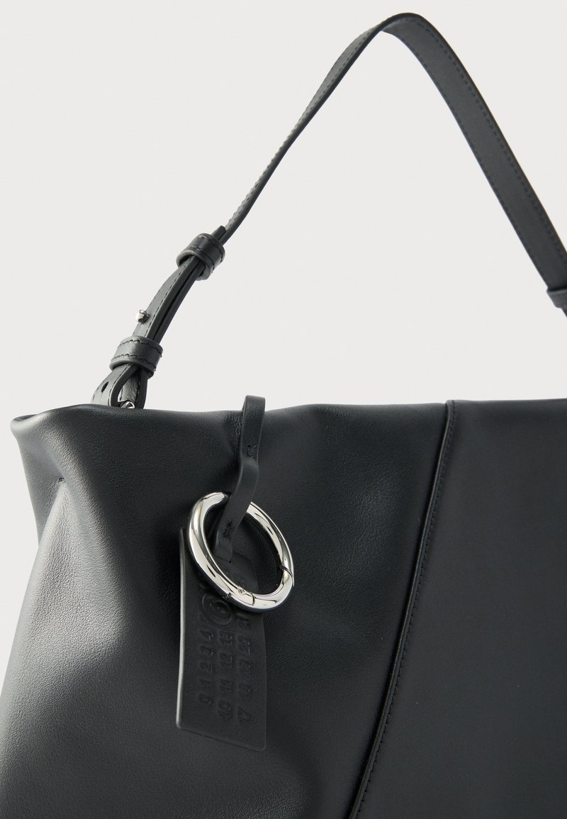 Schwarze Lederhandtasche mit einem einzelnen verstellbaren Riemen und einem silbernen, runden Ring, der an einem kleinen Lederetikett befestigt ist.