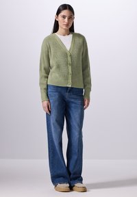 Ljusgrön cardigan med V-hals, ribbat mönster och fem knappar, tillsammans med lösa blåjeans och beige foderfleece-skor.